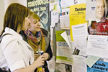 Zwei junge Frauen beim Zettelstudium an einem Schwarzen Brett einer Uni