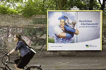 Radfahrerin fährt an einer Mauer mit Werbe-Plakat der Berliner Wasserbetriebe vorbei