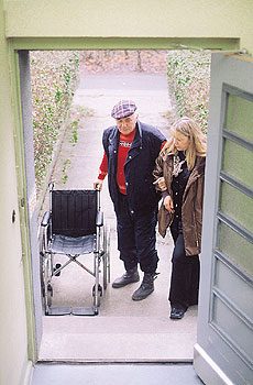 Senior mit Hilfsperson und Rollstuhl vor einer Haustür