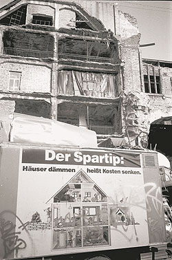Plakat mit der Aufschrift 'Der Spartip - Häuser dämmen heißt Kosten senken' vor einer Bauruine mit offener Fassade