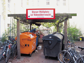 Ein Müllstandplatz mit diversen Müllbehältern, darüber ein großes Schild mit der Aufschrift 'Dieser Müllplatz ist Videoüberwacht, Die Hausverwaltung'