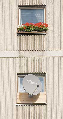 Kontraste: Plattenbaufassade mit zwei Fenstern; das obere ziert ein Blumenkasten, darunter ein Fenster mit einer auf einem Holzbrett befestigten Parabolschüssel