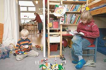 Zwei Kinder in ihrem offenen Kinderzimmer, im Hintergrund bereitet eine Frau Essen zu