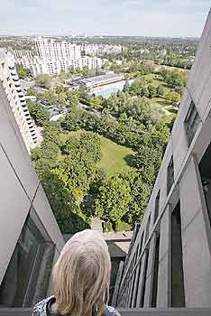 Ein Kind schaut aus einem oberen Fenster eines Hochhauses auf einen weit unten liegenden Park