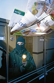 Ein Einbrecher mit Taschenlampe spiegelt sich im Briefkasten, der mit Zeitungen vollgestopft ist