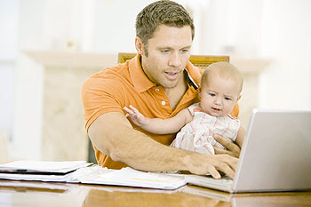 Junger Vater mit Kind auf dem Schoß vor seinem Laptop