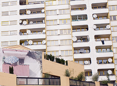 Kontrast: Balkonidylle mit Pflanzen und Sonnenschirm - aufgemalt auf ein Plakat - vor einer Betonfassade mit Fensterausschnitten und Parabolantennen