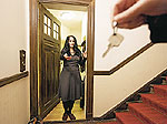 Eine Frau steht in geöffneter Wohnungstür mit ausgestreckter Hand nach einem Schlüssel, den ein Dritter in seiner Hand hält
