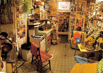 Eine Küche, vollgestellt mit Möbeln; an Wänden und Schranktüren eine Vielzahl von Karten, Stickern etc.