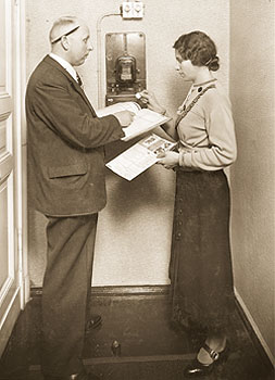 Ein Kassierer mit Stift über dem Ohr und Merkbuch in der Hand kassiert bei einer Frau den 'Stromgroschen' bar ab