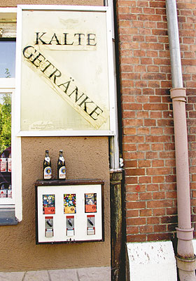 Schild an einer Hauswand mit der Aufschrift 'Kalte Getränke' über einem Süßigkeitautomaten, auf dem leere Bierflaschen stehen