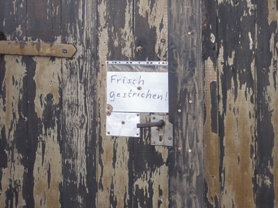 Alte Holzwand mit Tür, stark gemasert und verwittert mit einem Zettel mit der Aufschrift 'Frisch gestrichen'