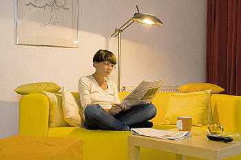 Frau sitzt auf einem Sofa und liest das Mietermagazin im Licht einer Halogenstehlampe