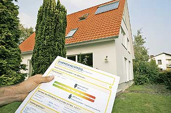 Hand mit einen Gebäude-Energieausweis vor einem Einfamilienhaus im Hintergrund
