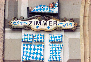 Schild an bayerischem Wohnhaus mit der Aufschrift 'Zimmer', ausgeschmückt mit einem Bett mit blauweißer Bettdecke sowie säuberlich ausgelegter blauweißer Doppelbett- und Einfachbettausstattung
