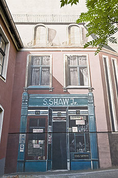 Fassadenmalerei simuliert Architektur: Ladengeschäft und Fenster auf 1. + 2. Stock auf einer kahlen Wand