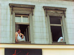Altbaufassade mit zwei offenen Fenstern; im einen Fenster ein dickbäuchiger Mann mit Bierdose in der Hand, er schaut auf eine Frau mit Kopftuch, die aus dem anderen Fenster nach unten schaut