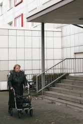 Frau mit Rollator vor einer Hauseingangs-Treppe