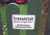 Schild am Gartenzaun mit der Aufschrift: Irrenanstalt - Betreten auf eigene gefahr! Mit Belästigungen muß gerechnet werden!