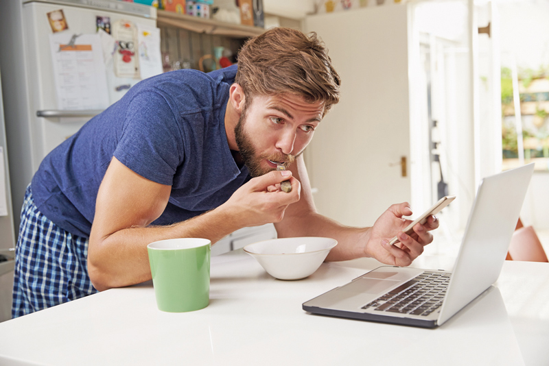 Mann mit Smartphone und Laptop beim Frühstück