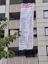 Das Foto zeigt einen simplen Wohnblock mit inneliegenden Balkonen; vor drei übereinander liegenden Balkonen hängt ein Stoffbanner mit allen Mängeln in der Adler Siedlung in Lichtenrade.