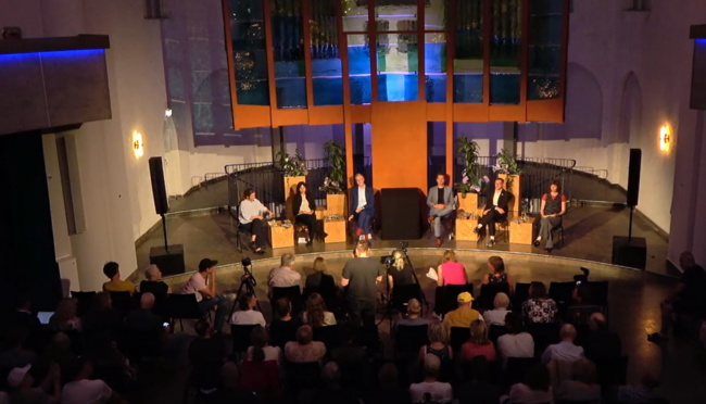 Wahlforum Snippet 2 Das Foto zeigt aus der Schrägperspektive von oben das Altarinsel der Genezarethkirche in Berlin Neukölln. Vier Spitzenkandidat:innen von CDU, SPD, Grüne und Linke sitzen auf dem Podium. Im Vordergrund ist das Publikum zu sehen.